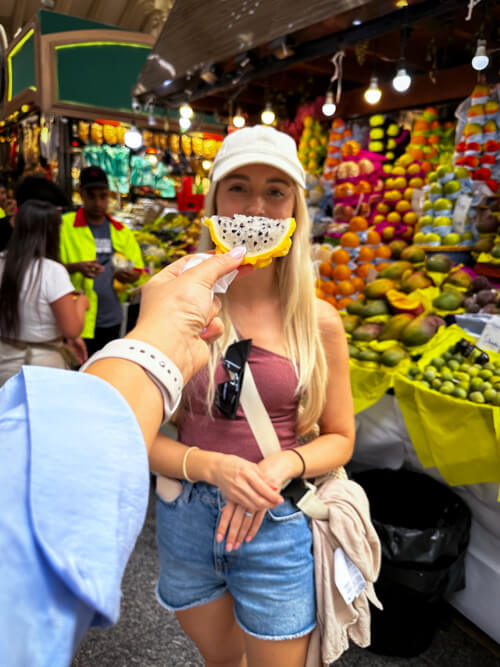 Me posing with a slice of yellow dragonfruit in front of a fruit stand.