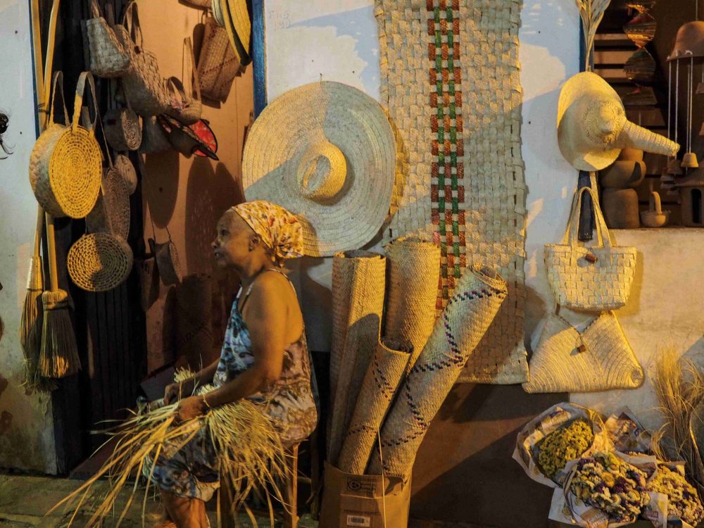 A Brazilian woman sitting on a stool in front of a handicraft shop and making a basket out of straw 