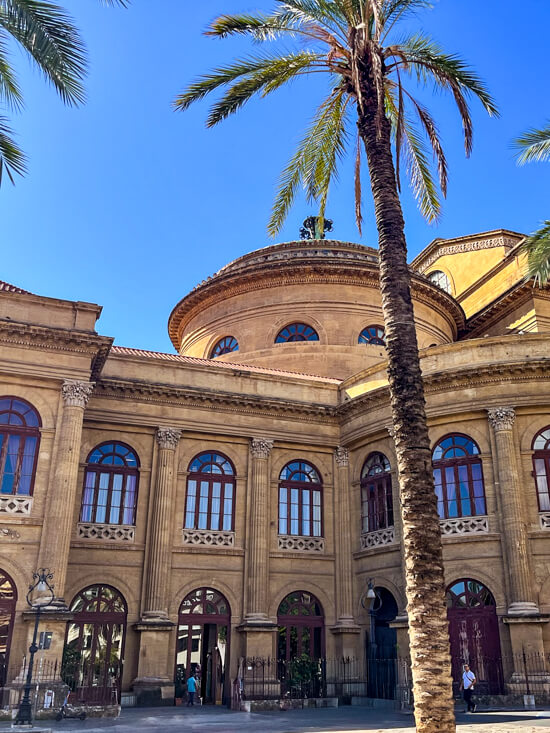 A tall palm tree in front of the beige stone facade of Teatro Massimo on a clear sunny day.