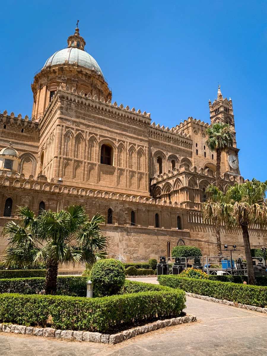 Palermo Cathedral with its intricate beige facade featuring elements from various architectural styles. In front of the cathedral there's a small park with shrubs, low palm trees and walking paths.