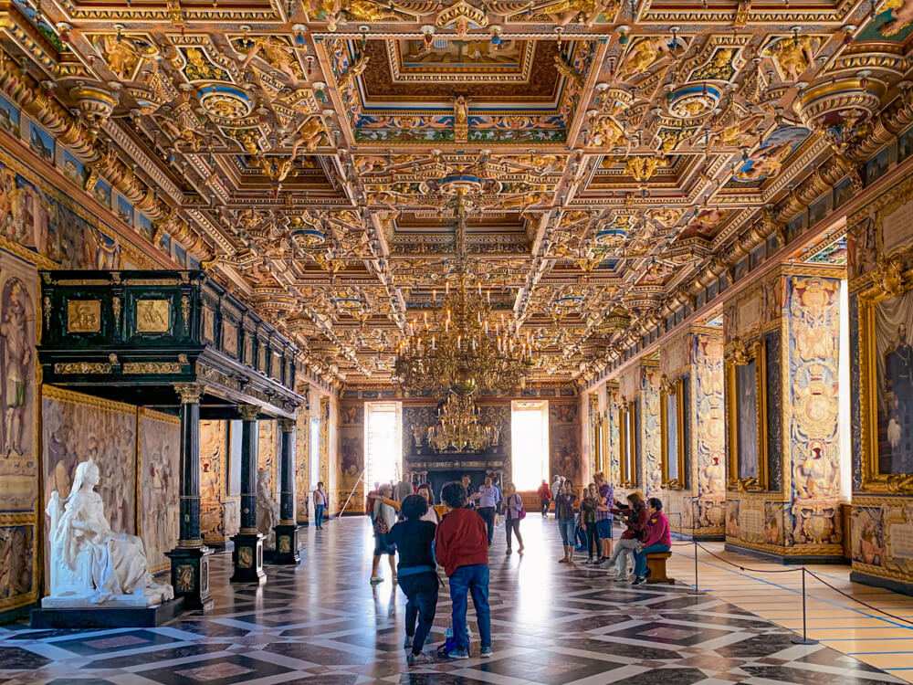 The ornate Great Hall of Frederiksborg Castle in Denmark, featuring a ceiling decorated with golden details, crystal chandelier, marble sculptures, and a geometric marble floor with tourists exploring the space.