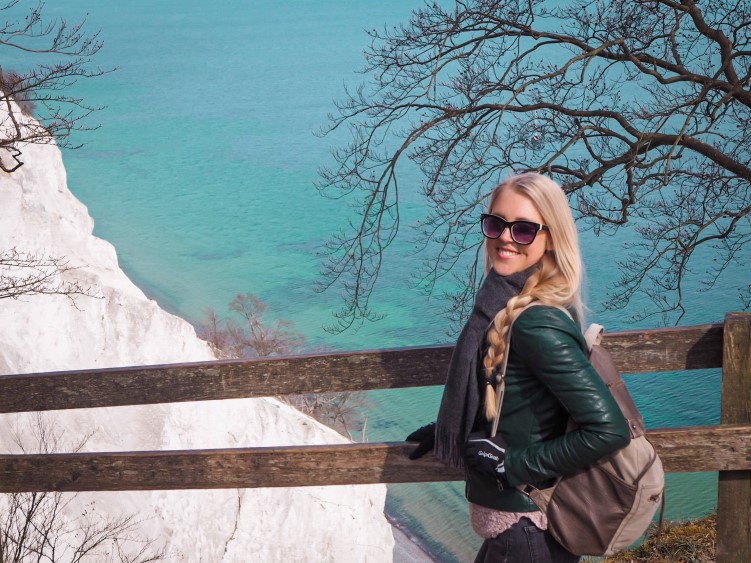 Me standing next to a railing with a view of bright blue water and white chalk cliffs of Møn in the background.