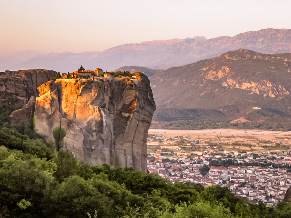 A view of The Holy Trinity Monastery perched on top of a steep vertical boulder in Meteora during sunrise. The town of Kalambaka is visible down below.