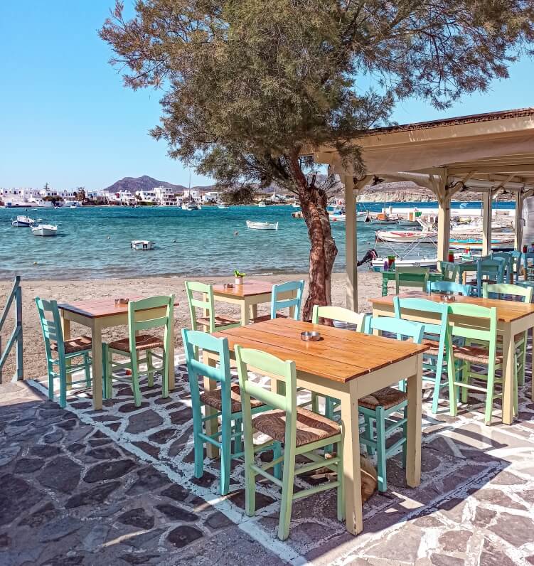 wooden tables and pastel-colored chairs at a beachfront cafe overlooking the sea in the village of Pollonia in Milos