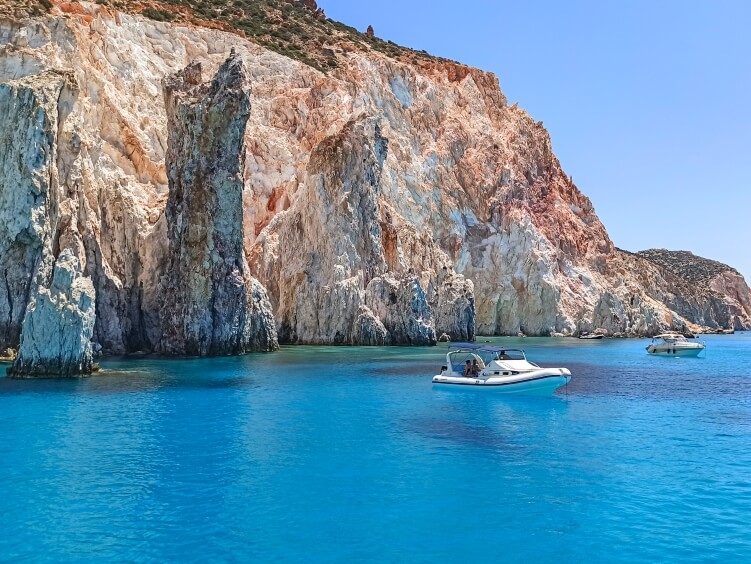 A white boat floating in vivid blue sea, surrounded by cliffs in shades of orange and white at Polyaigos Island in the Aegean Sea, Greece