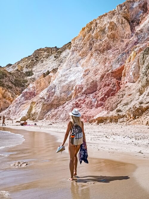 Me walking along the sandy shore of Firiplaka Beach with mountains in shades of red, white and orange in the background
