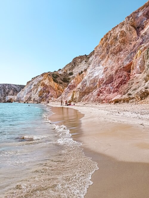 Gentle waves along the sandy Firiplaka Beach bordered by volcanic mountains in different shades of oranges and red