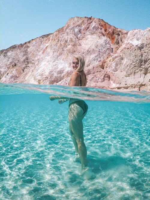 Me standing waist-deep in the crystal clear sea at Firiplaka Beach with colorful cliffs behind me