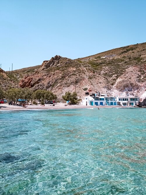 A bay with calm blue water, surrounded by a hilly landscape and a few white boat houses at Firopotamos village