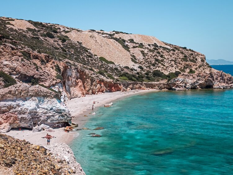 A small secluded cove with turquoise water, a white pebble beach and orange  hills around it at Kastanas Bay