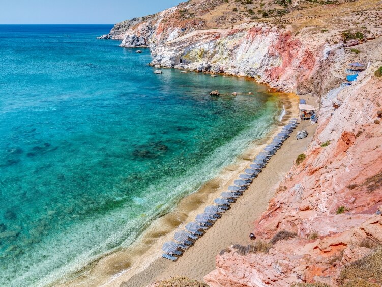 Emerald water and umbrellas on a stretch of golden sand at Paleochori Beach bordered by cliffs in shades of red and orange