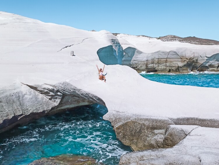 Me sitting in the middle of a white rock arch extending over a natural pool full of turquoise water at Sarakiniko Beach