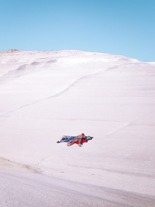 a man sunbathing on a red beach towel on the white smooth rock surface of Sarakiniko Beach