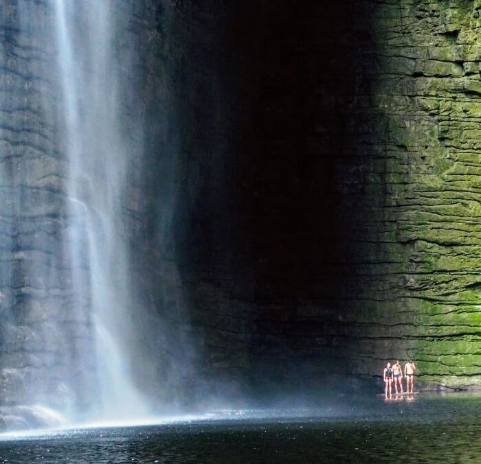 Three people standing next to Cachoeira da Fumacinha waterfall surrounded by a dark canyon