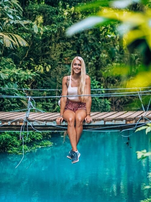 a woman sitting on a wooden bridge above the bright blue water of Rio Celeste in Costa Rica