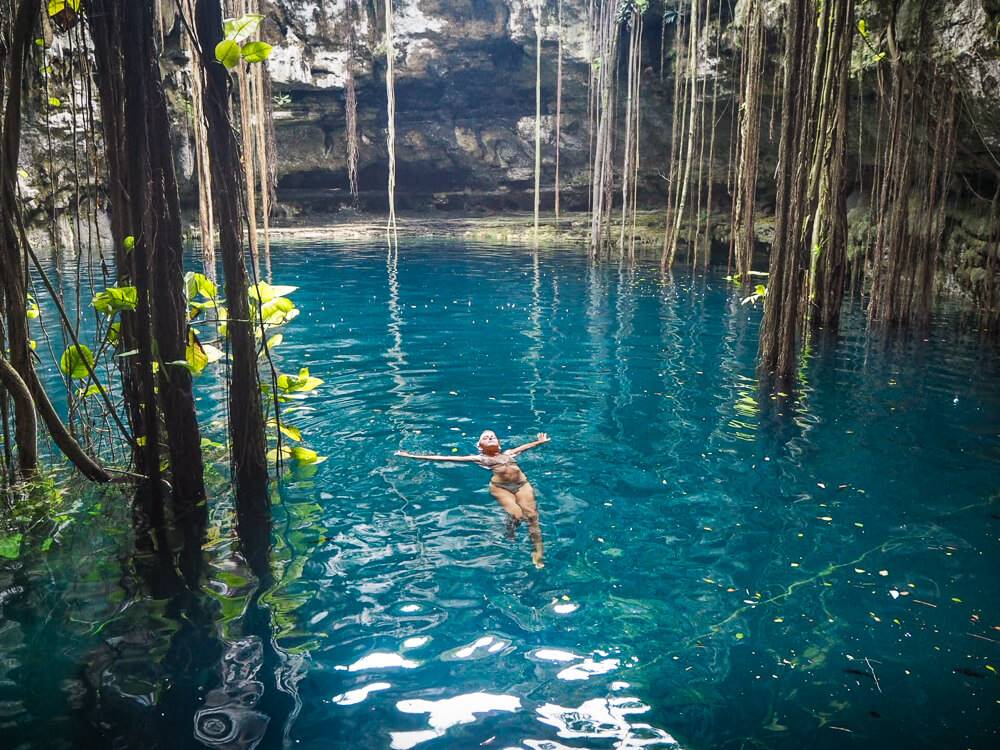 a girl swimming in blue water between hanging vines in Cenote Oxman in Mexico, one of the most unique things to do in Valladolid