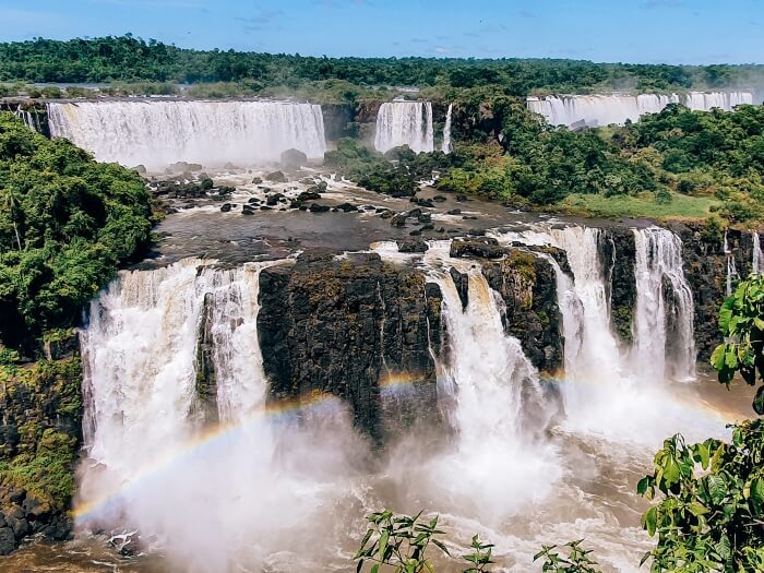 Lush vegetation and a rainbow in front of the white cascades of Iguazu Falls viewed from the Brazilian side