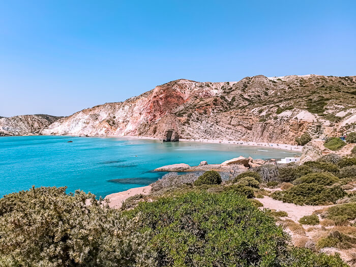 A bay with turquoise water, surrounded by green vegetation and orange mountains at Firiplaka Beach