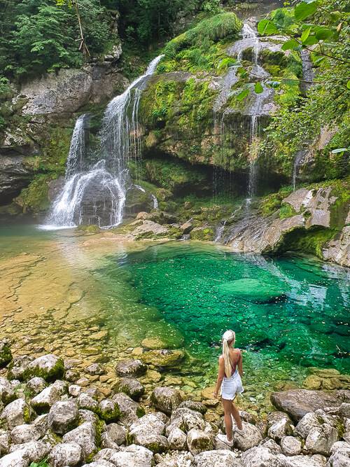 a woman standing on rocks in front of a blue-green basin and Virje Waterfall, one of the best waterfalls in Slovenia