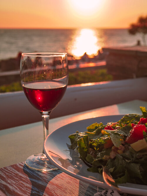 a salad and a glass of red wine with a backdrop of a sunset at a restaurant in Antiparos Island