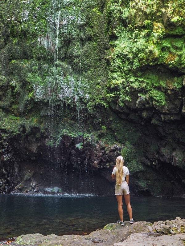 Me standing at the edge of a pool of water gazing up at the Caldeirão Verde waterfall trickling down a moss-covered rock face.