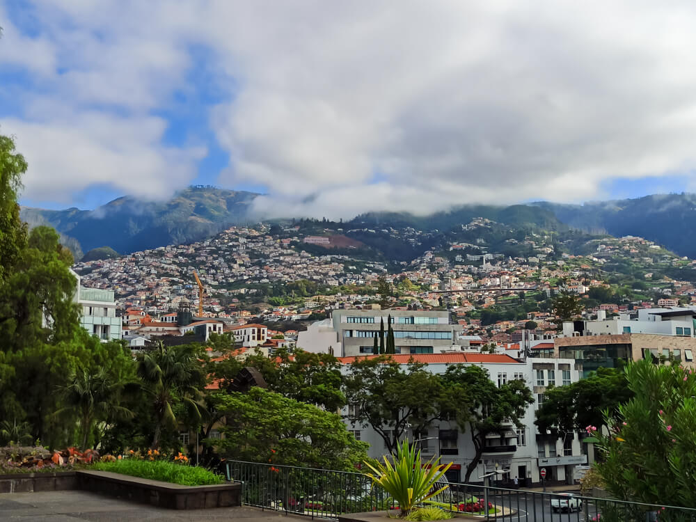 Panoramic view of Funchal, Madeira, with white, terracotta-roofed buildings on the hillside, framed by green mountains under a partially cloudy sky.