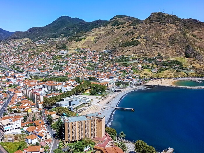 Panoramic view of the coastal town of Machico, dotted with houses with terracotta roofs and surrounded by green mountains.