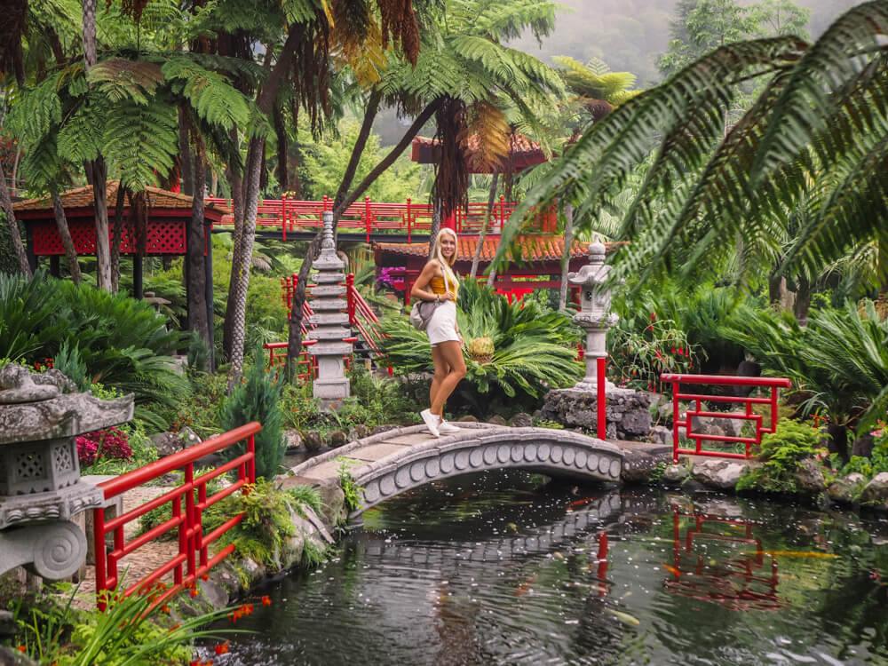 Me standing on a small arched bridge over a koi pond surrounded by lush tropical greenery and small pagodas at Monte Palace Tropical Garden, Madeira.