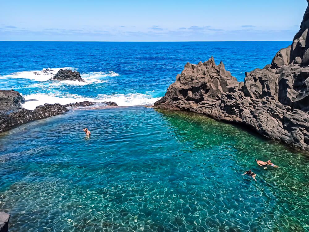 Swimmers in the clear turquoise natural pools at Seixal, Madeira, framed by jagged volcanic rocks and the open ocean behind them.