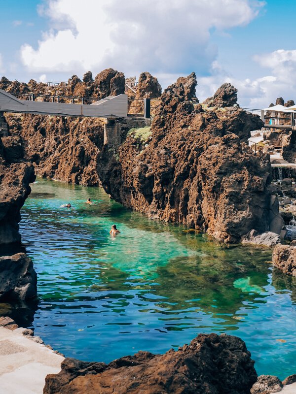 Swimmers in the crystal-clear emerald waters of Porto Moniz natural pools surrounded by rugged brown volcanic rocks.