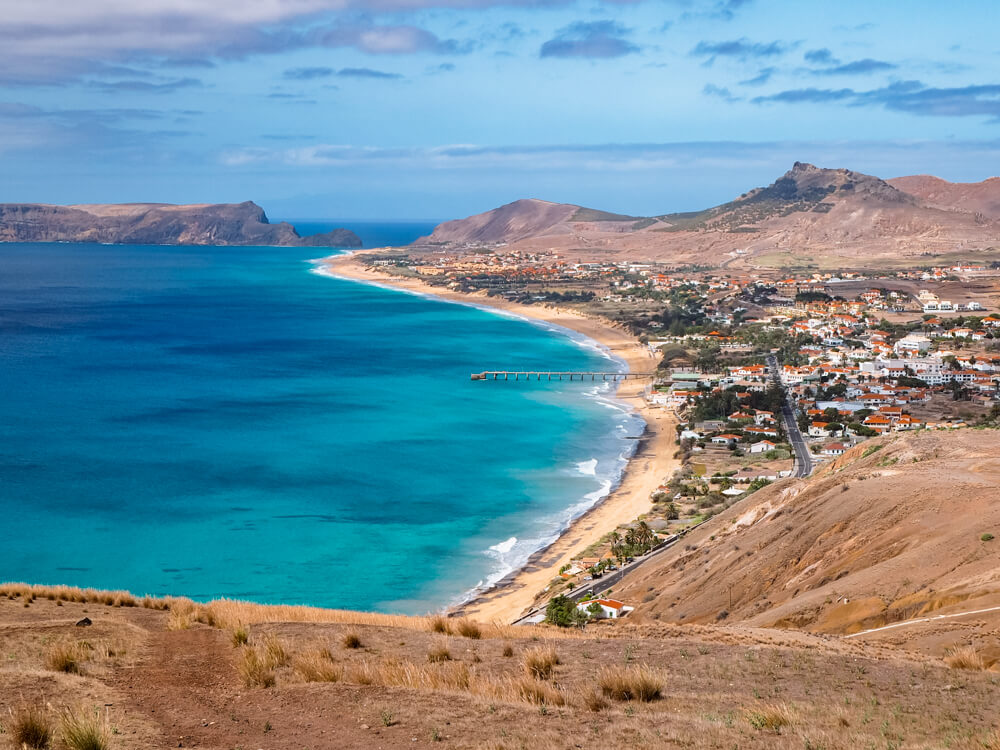 A panoramic view of the coastline of Porto Santo Island in Madeira with its dry terrain and a long sandy beach with golden sand and vivid turquoise water.