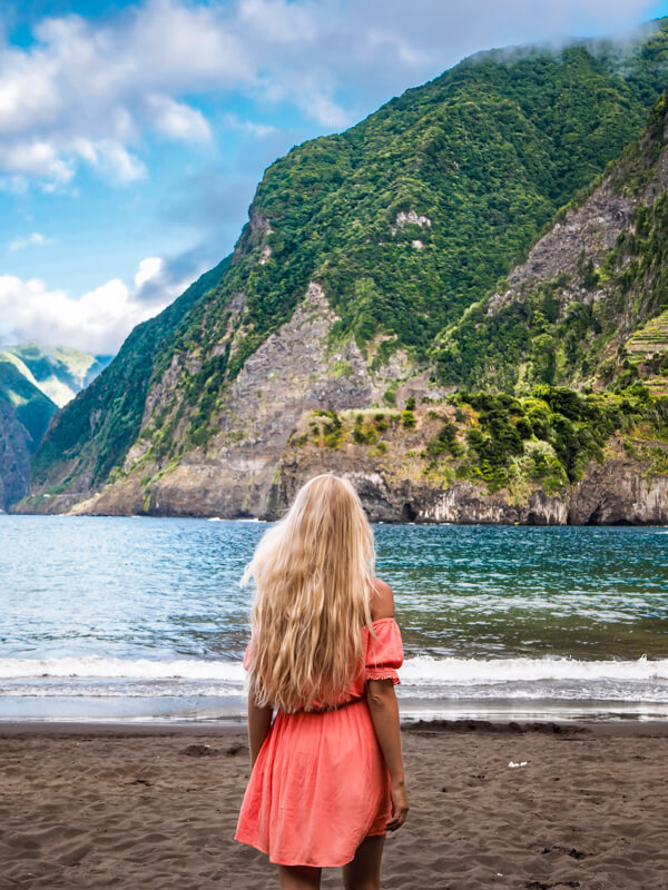 Me in a coral dress walking on the black sand beach of Seixal, Madeira, with the steep green mountains rising from the ocean in the background.