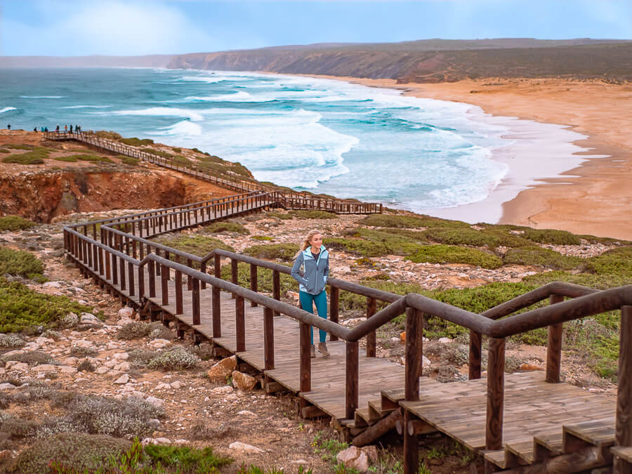 A woman walking on a boardwalk overlooking the huge Praia da Bordeira, a popular beach among surfers