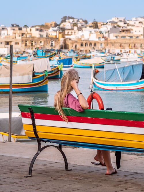 a woman sitting on a colorful bench at the harbor of Marsaxlokk fishing village