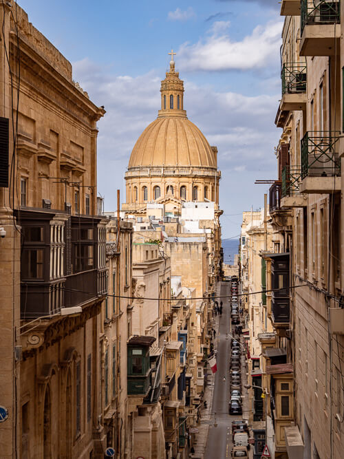 a hilly street with a view of the big dome of the Basilica of Our Lady of Mount Carmel in Valletta
