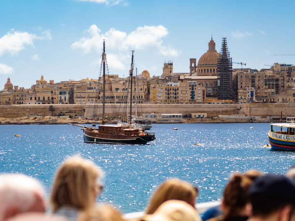 An old wooden sail boat with the skyline of Valletta in the background on a sunny day, viewed from a ferry to Sliema.