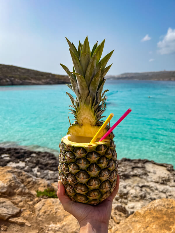 My hand holding a cocktail served in a pineapple. In the background is the clear turquoise water of the Blue Lagoon.