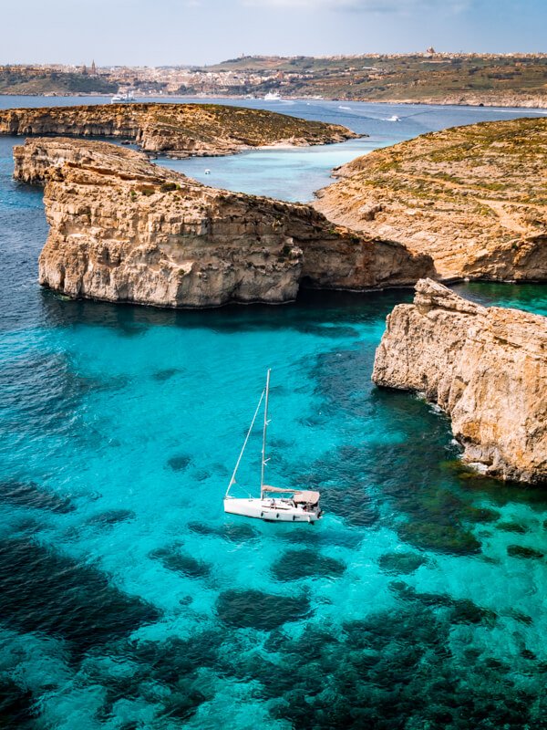 A white sailing boat surrounded by vibrant blue water and steep cliffs at Crystal Lagoon on Comino Island in Malta.