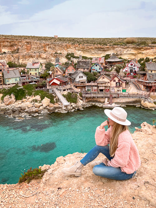 A woman sitting on a cliff overlooking the Popeye Village theme park, one of the most famous Malta Instagram spots