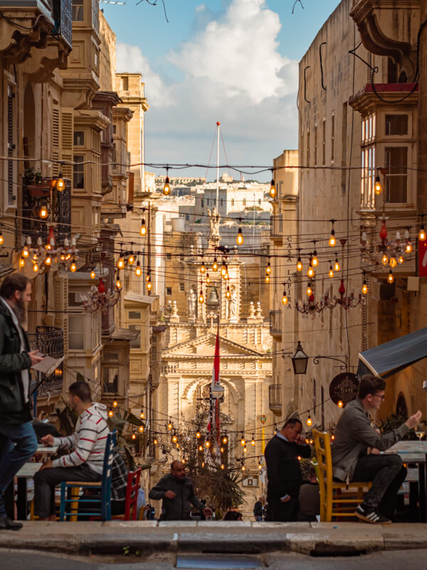 People dining on the steps of the sloped St. Lucia's Street, surrounded by beige stone buildings in Valletta.