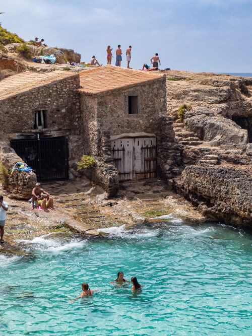People swimming in the small bay of Cala S'Almunia next to old boat houses.