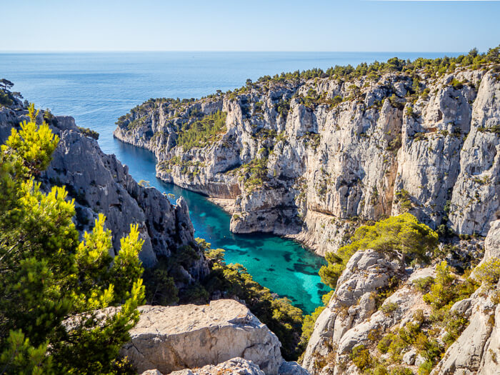 A view over the neon blue water of the narrow Calanque d'En Vau bay surrounded by limestone cliffs.