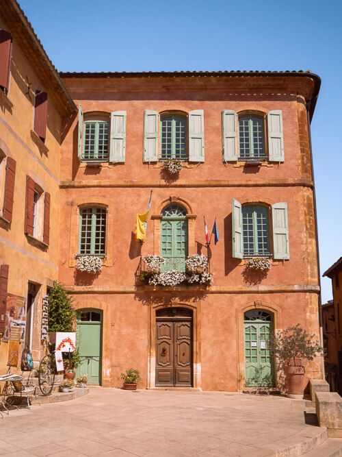 An ochre-colored house with blue window shutters in the village of Roussillon, a must-visit spot on every South of France road trip.