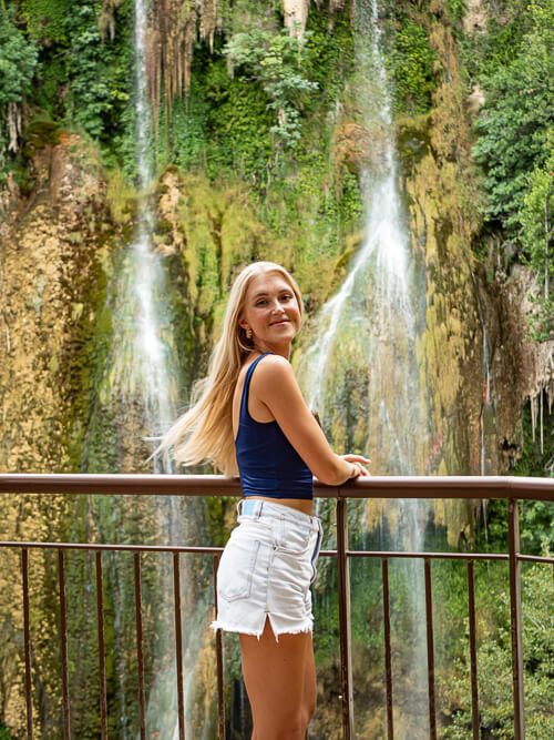 A woman leaning on a railing with the Cascade de Sillans waterfall and its lush vegetation as a backdrop