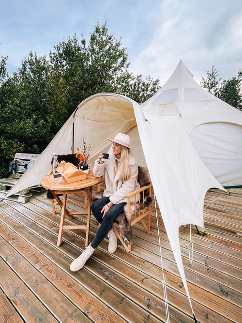 A woman sitting on a chair in front of a white glamping tent, sipping a cup of coffee