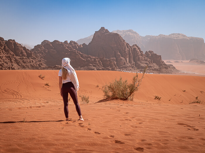 A woman standing in orange sand with a backdrop of rugged sandstone mountains in the Wadi Rum desert