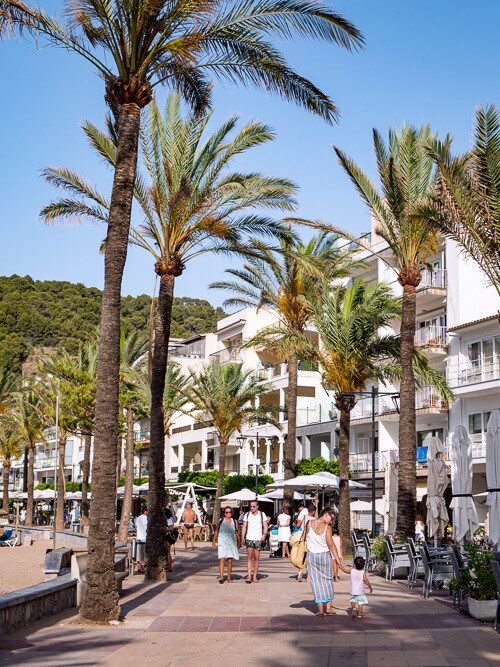 Tourists strolling on a palm-tree-lined promenade in Port de Soller on a sunny afternoon with blue skies.