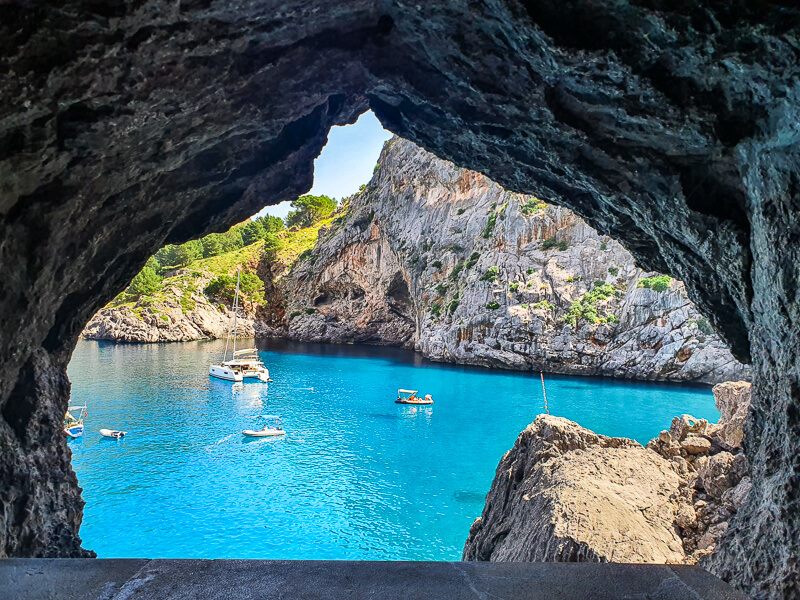 A natural window inside a rock tunnel overlooking a bay with clear turquoise water at Sa Calobra, Mallorca.