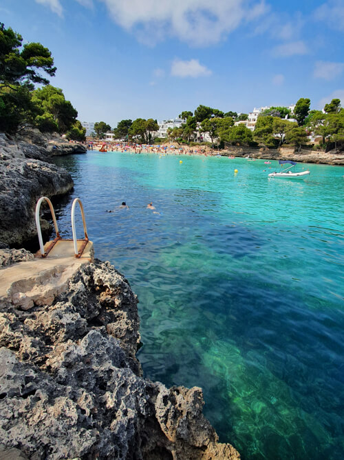 A ladder descending into the water at the rocky shore of the Cala Gran beach with its vivid turquoise water.