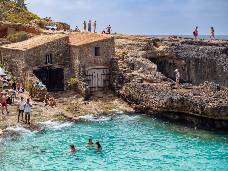 People swimming in the small sheltered bay of Cala S'Almunia next to old boat houses and rocky shores